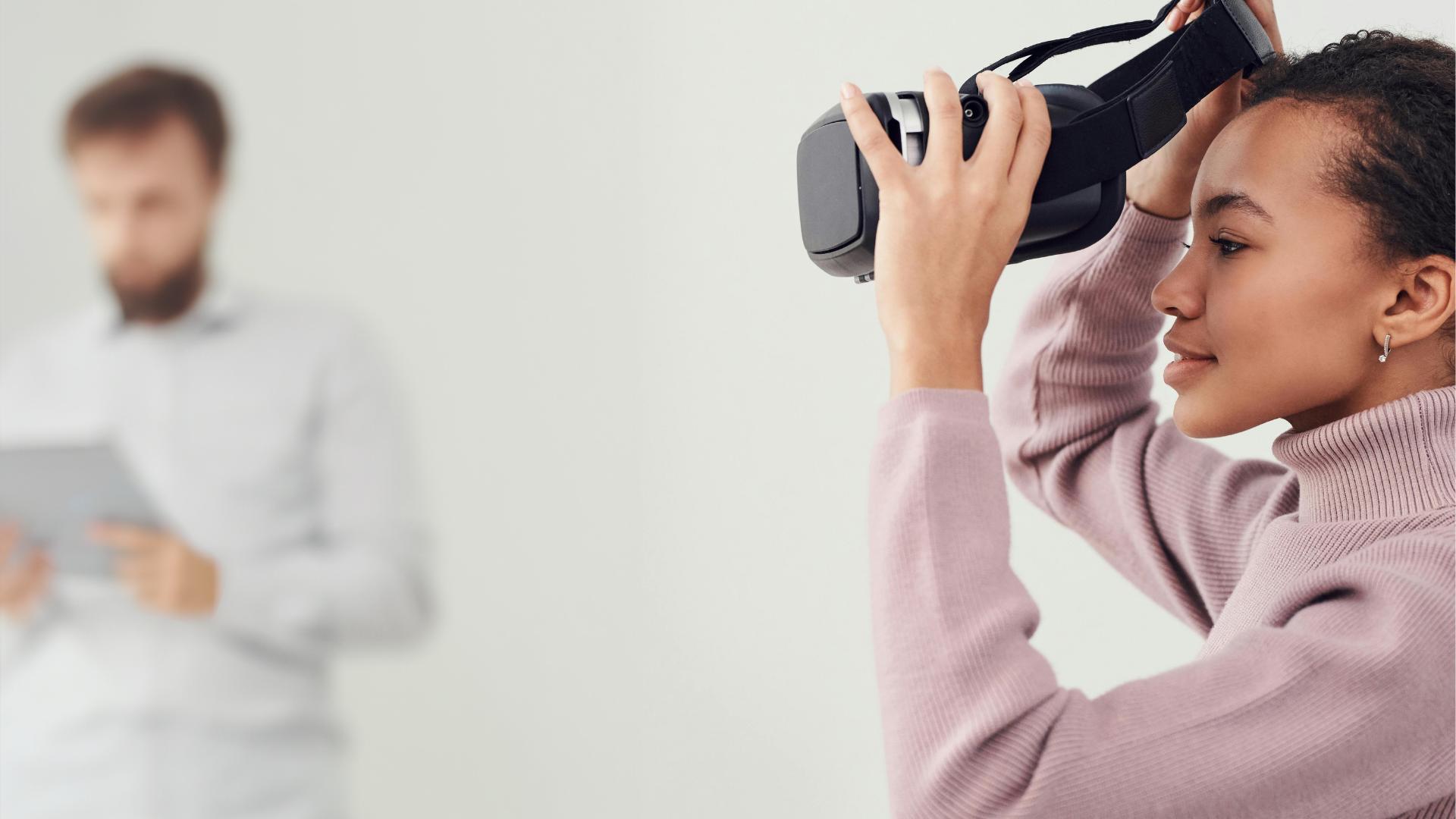 One person puts on a VR headset while another person in the background uses a tablet.